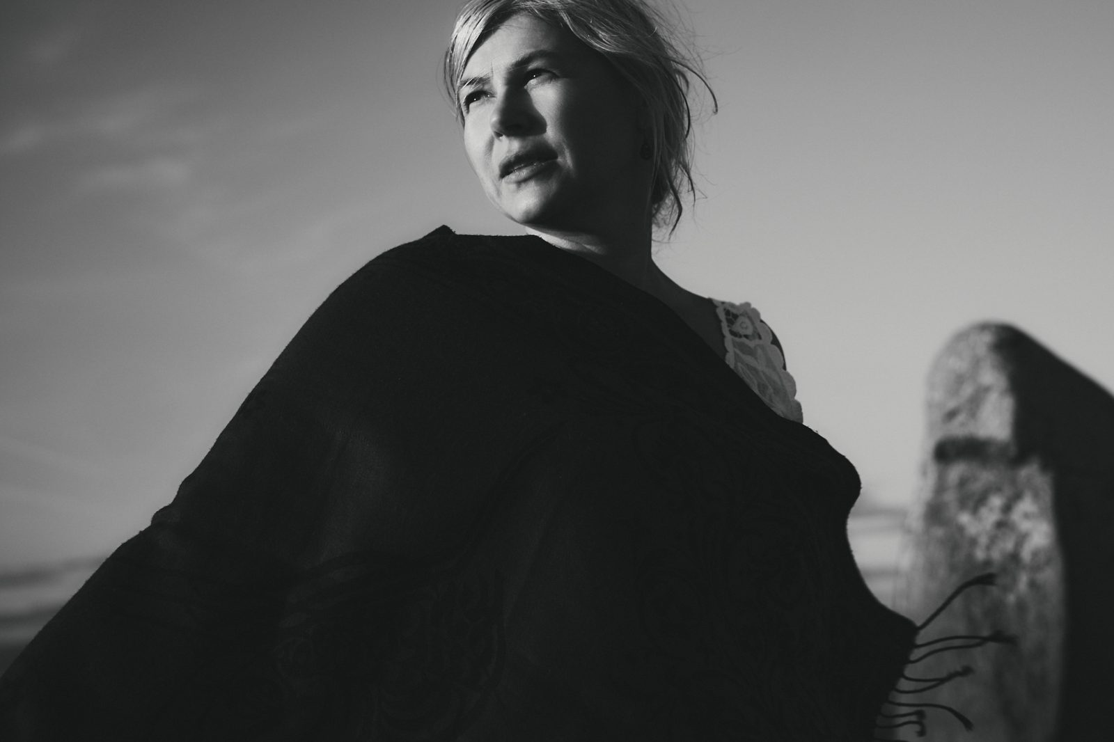Woman in a dark shawl beneath open sky beside a standing stone, black and white portrait photography, Ale’s Stenar, Kåseberga, Sweden, 2026